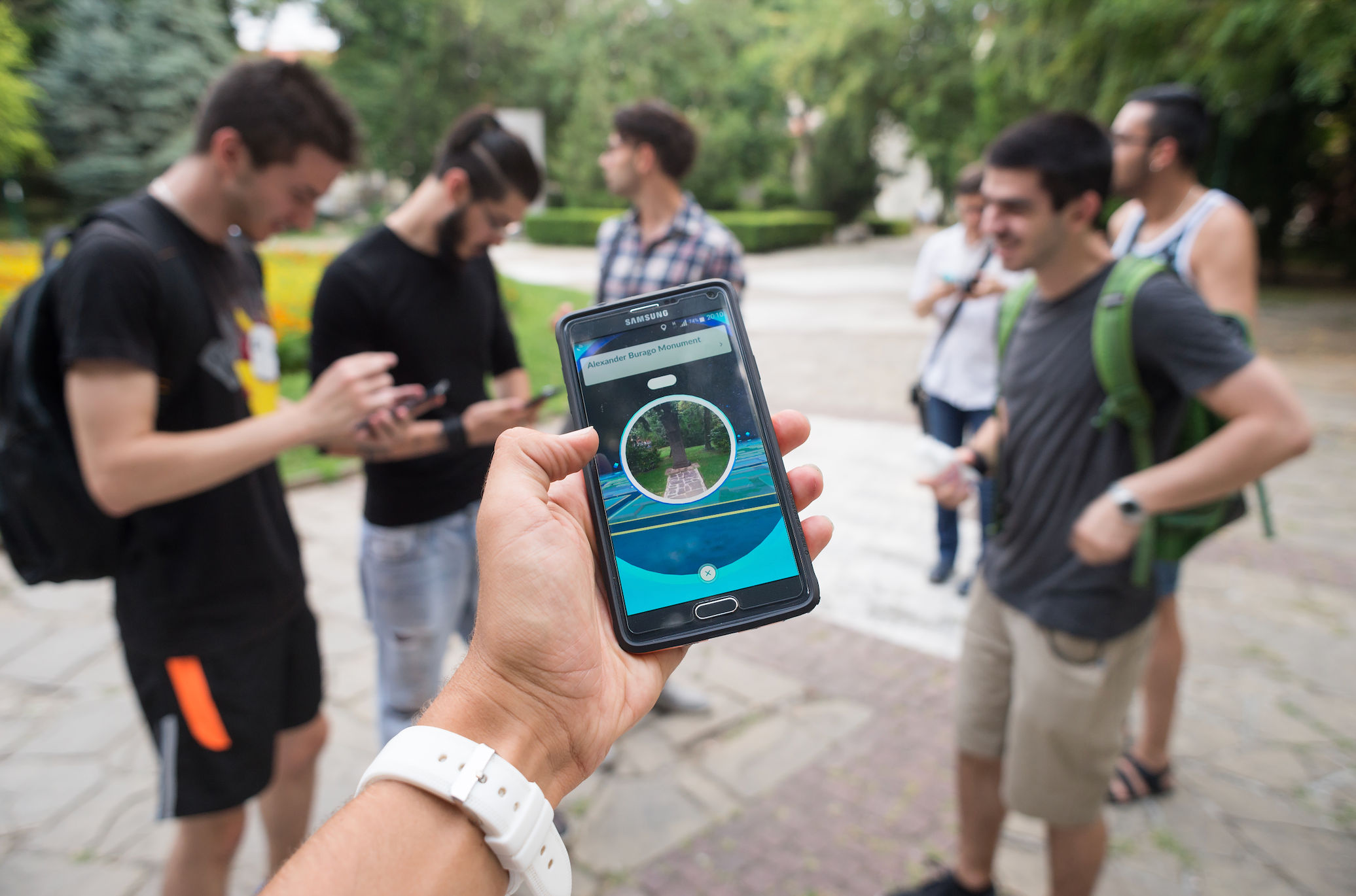 A photo of someone's arm extended with their mobile phone in their hand. They're playing the augmented reality game Pokémon GO. In the background you can see a group of six people, also playing Pokémon GO as demonstrated by them looking at their phones. They're standing in a park.